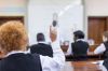 Waist up shot of African male teacher leading biology class, out of focus, students foregrounded with hands up, Cape Town, South Africa