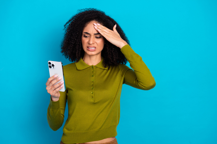 Young woman with curly hair looking at smartphone against blue background showing frustration