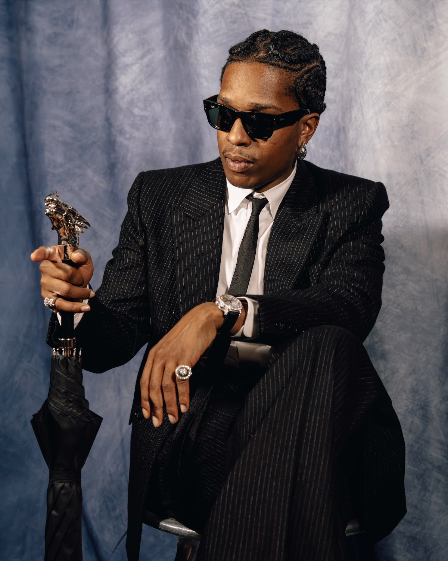 A well-dressed man in a pinstriped suit and sunglasses holding a trophy-like object, seated against a blue backdrop.