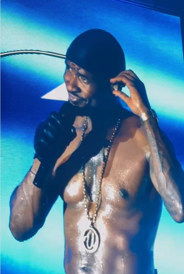 Shirtless male performer on stage wearing jewelry and a black jacket, with a microphone in hand.